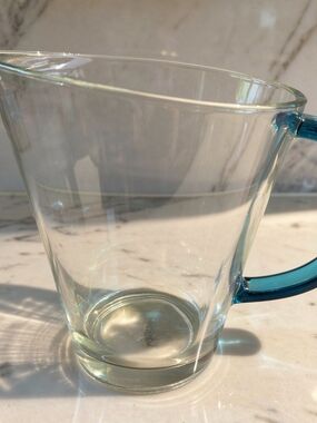 A clear glass pitcher with a blue handle by Pasabahce